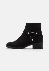 Botines de ante negro hasta el tobillo con punta redondeada, tacón bajo en bloque y una correa ajustable con herrajes metálicos y botones decorativos.