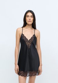 Sort satin camisole med justerbare stropper, med en dyp V-hals kantet med blonder og en bølget blondekant. Jevn tekstur generelt.