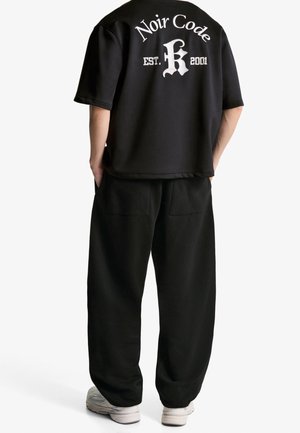 Person iført løse sorte bukser, en sort kortærmet skjorte med teksten "Noir Code EST. 2001" på ryggen og hvide sneakers, står med hænderne i lommerne.