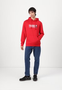 Felpa rossa con logo bianco "TOMMY" e piccolo design della bandiera, dotata di tasca frontale e cappuccio con coulisse, abbinata a jeans blu e sneaker nere.