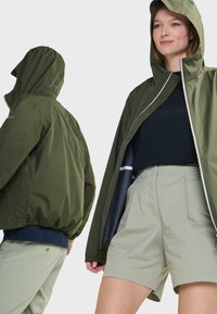 Olijfgroene, met rits gesloten jas met een kap. Lichtgewicht materiaal en elastische manchetten. Gecombineerd met lichtgekleurde khaki short, met een ontspannen pasvorm.