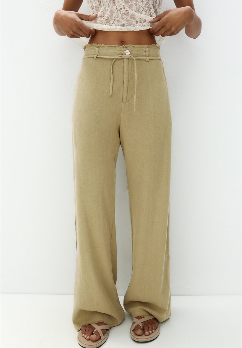 Beige wijde broek met een drawstring taille, knoopdetail en rafelige zoom, gemaakt van een lichte, gestructureerde stof. Gepaard met sandalen.