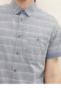 Camisa de manga corta con botones en gris claro con un patrón de cuadros. Cuenta con un bolsillo en el pecho y detalles en contraste azul en los puños.