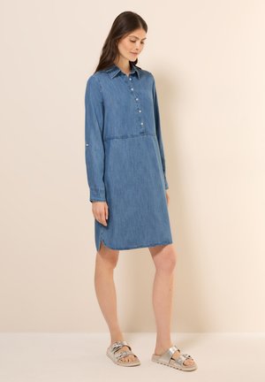 Femme portant une robe en jean bleue à manches longues avec boutons et des sandales argentées, debout devant un fond uni beige clair.
