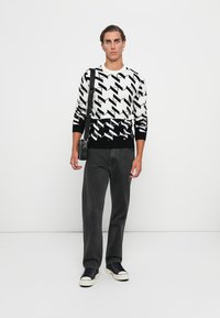 Schwarzer und weißer gemusterter Pullover mit V-Ausschnitt, der geometrische Designs aufweist. Kombiniert mit dunkelgrauen Jeans und schwarzen Sneakers.