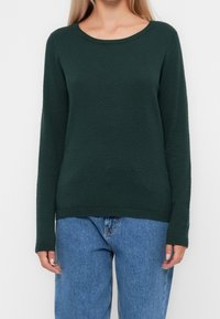 Mörkgrön texturerad tröja med rund halsringning, långa ärmar och ribbad nederkant, tillsammans med blå jeans.