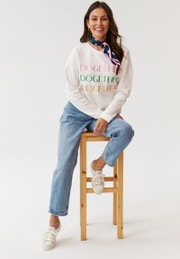 Felpa bianca con il testo multicolore "DOGETHER", abbinata a jeans blu chiaro e sneakers bianche. Una sciarpa blu con motivi a cuore completa il look.