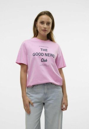 T-shirt en coton rose à manches courtes, avec un texte imprimé en noir : "NOUS VOUS PRÉSENTONS LA BONNE NOUVELLE Club" accompagné d'un design graphique.
