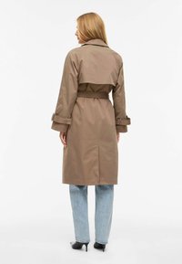 Trench-coat beige avec une taille ceinturée, un large col et des manches longues. Comprend une fente arrière et des poignets retroussés. Associé à un jean bleu clair.