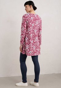 Rote Blumentunika mit weißen und blauen Blumenmustern, langen Ärmeln, einem runden Ausschnitt und einem leicht ausgestellt geschnittenen Saum, kombiniert mit dunkelblauen Leggings.