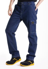 Pantalons cargo en denim bleu foncé avec des coutures blanches contrastantes, dotés de poches latérales et d'un badge logo. Portés avec des chaussures noires et jaunes.