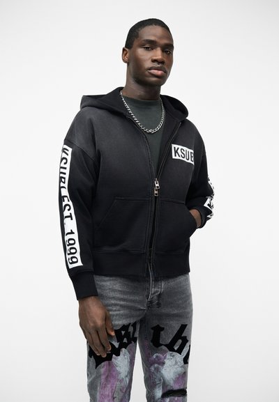 Νεαρός άντρας που φοράει μαύρο φούτερ με φερμουάρ και λευκό κείμενο "KSUB" καθώς και "KSUB EST. 1999" στα μανίκια, συνδυασμένο με γκρι τζιν με τύπωμα και ασημένιο αλυσίδα λαιμού.