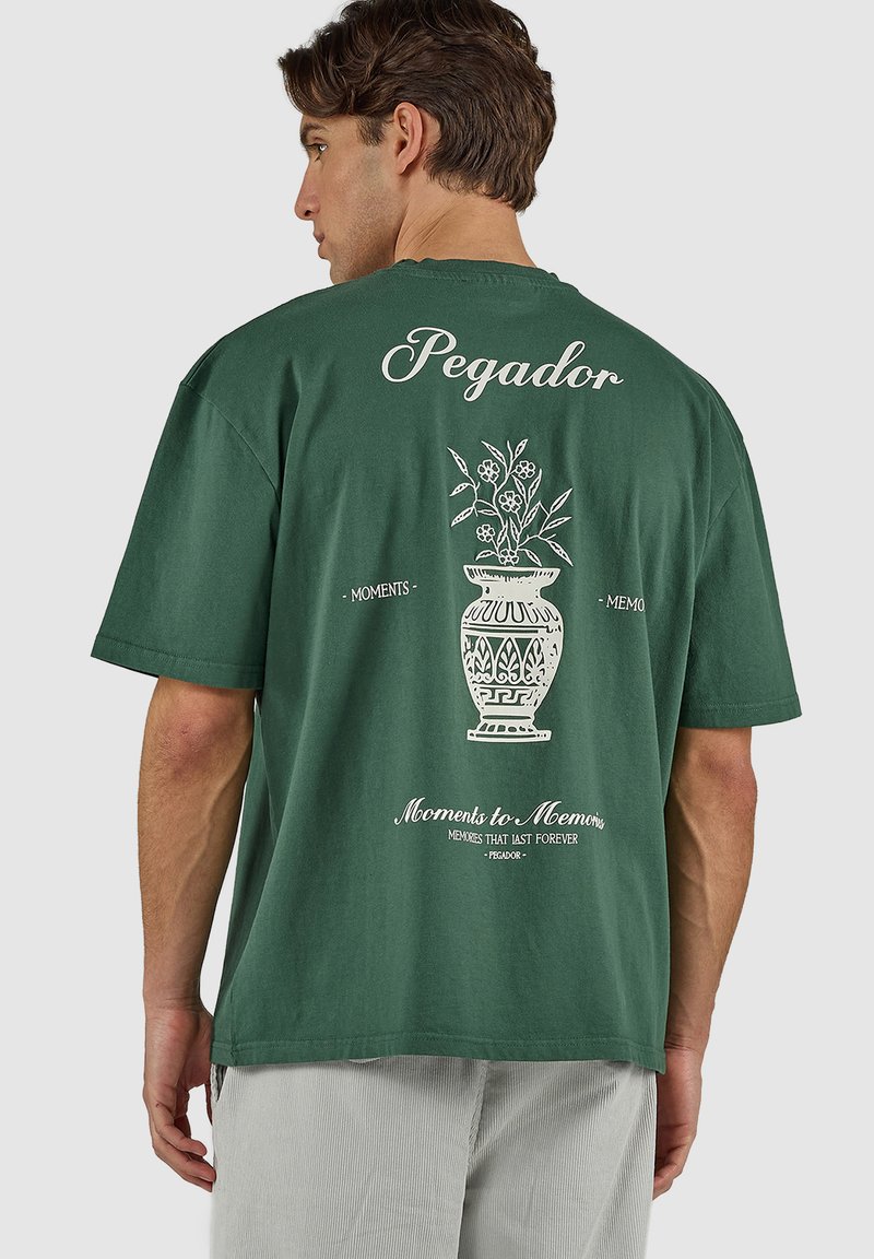 Mężczyzna noszący zieloną, oversize'ową koszulkę z białą ilustracją wazy z kwiatami oraz napisem "Pegador" i "Moments to Memories" na plecach.