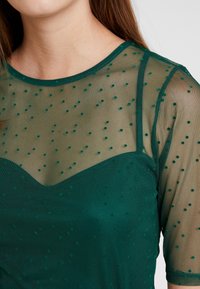 Top de malla verde con mangas cortas, con cuello redondo, patrón de puntos y superposición transparente sobre una capa base verde sólido.
