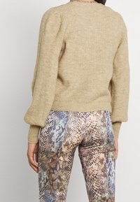 Suéter beige de punto con mangas abullonadas y textura acanalada. Combinado con pantalones ajustados de estampado de serpiente en tonos terrenales, mostrando intrincados detalles del patrón.