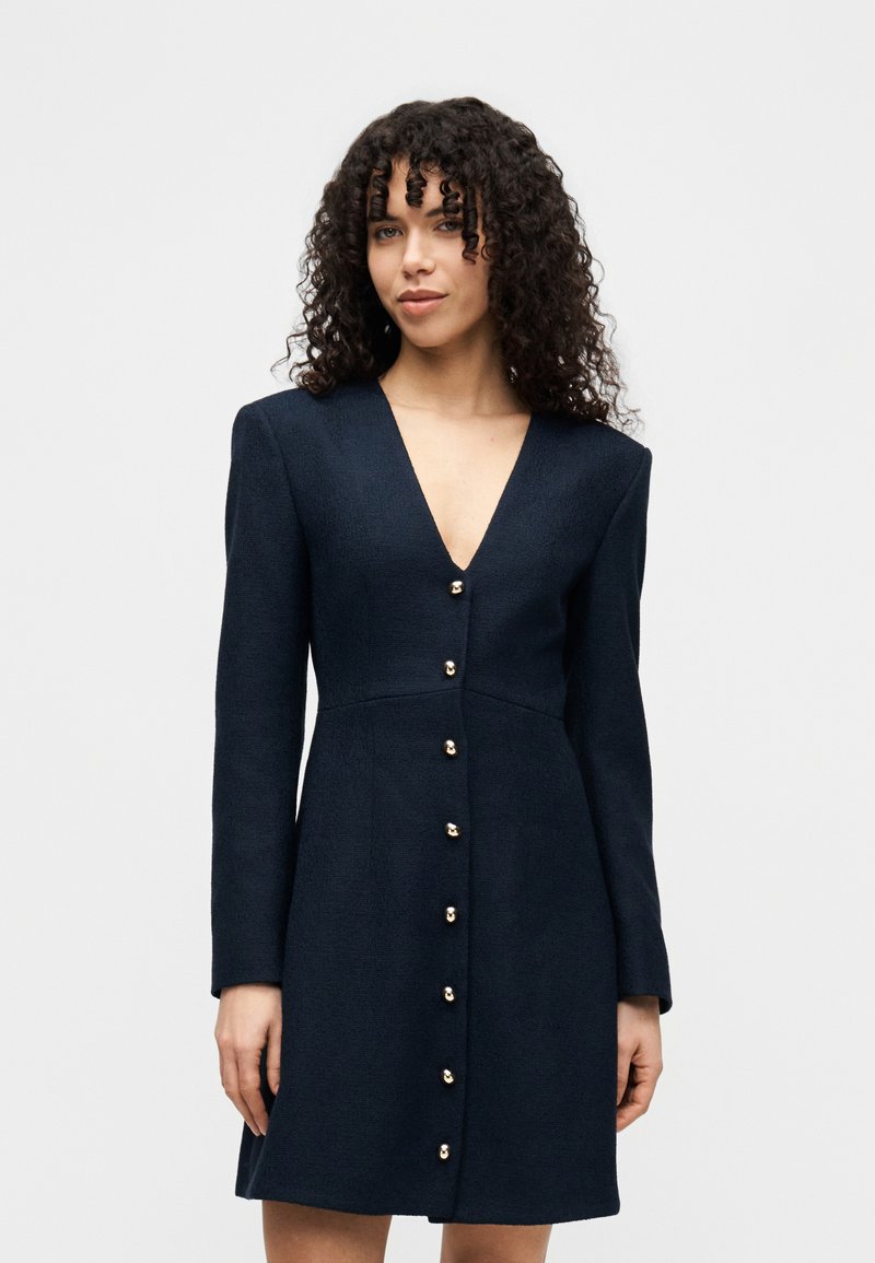Φόρεμα από μάλλινο μείγμα σε ναυτικό μπλε χρώμα, με βαθύ V-λαιμόκοψη, μακριά μανίκια και χρυσά κουμπιά μπροστά. Ραμμένη εφαρμογή με ελαφριά φούστα στο τελείωμα.