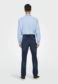 Hellblau gestreifte Langarmbluse, dunkelblaue Jeans und marineblaue Schuhe. Sichtbare Nähte, Knopfmanschetten und zwei Gesäßtaschen an der Jeans.