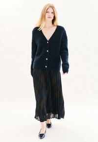 Cárdigan de punto azul marino con escote en V profundo, botones grandes y mangas largas, combinado con una falda de cuadro en negro y verde con pliegues y zapatos planos azul marino.