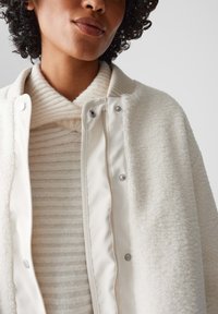 Crème fleece jack met een gladde crème binnenvoering, zilveren drukknoopsluitingen, gedragen over een licht gestreepte trui. Zachte textuur en gezellige ontwerp.