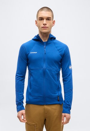 Noor mees, kes kannab sinist Mammuti lukuga kapuutsiga jopet ja pruune pükse, seisab ühevärvilise valge tausta ees.