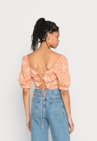 Blusa floral laranja com mangas bufantes e costas abertas, com detalhe de amarrar em renda. Usada com jeans de cintura alta azuis.