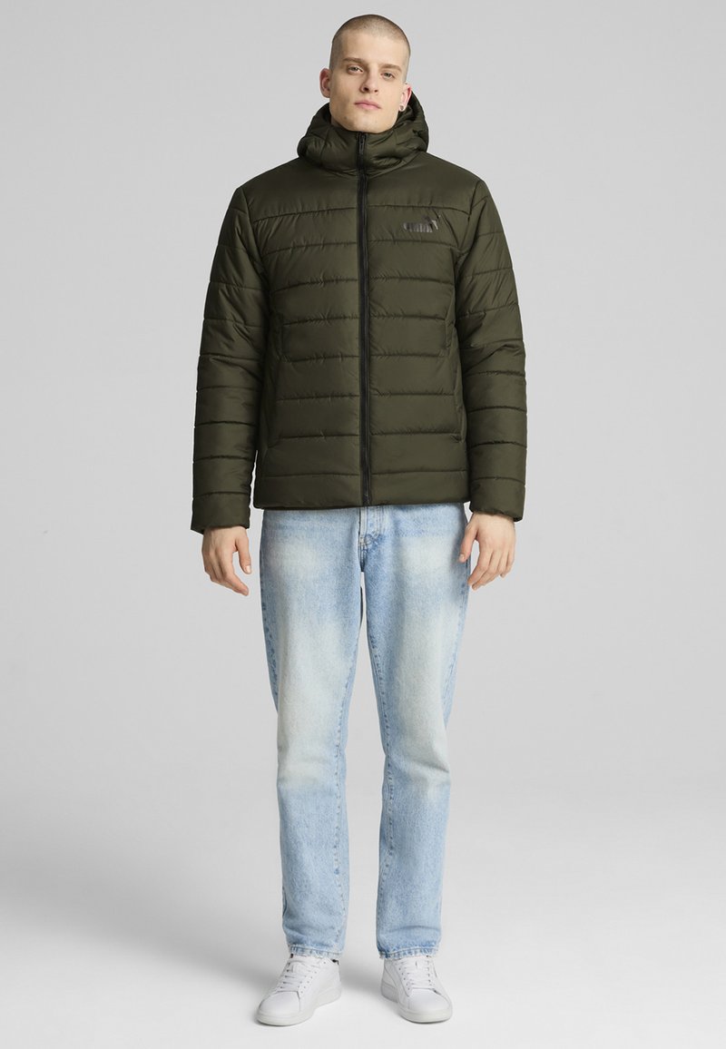 Giubbotto puffer verde oliva con chiusura a zip e cappuccio, caratterizzato da una texture trapuntata. Abbinato a jeans azzurri e sneakers bianche.