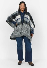 Rutig poncho i grå, svart och vit färg, med en triangulär snitt, mjuk textur och fransdetaljer vid kanterna. Bärs över en jeansskjorta och jeans.