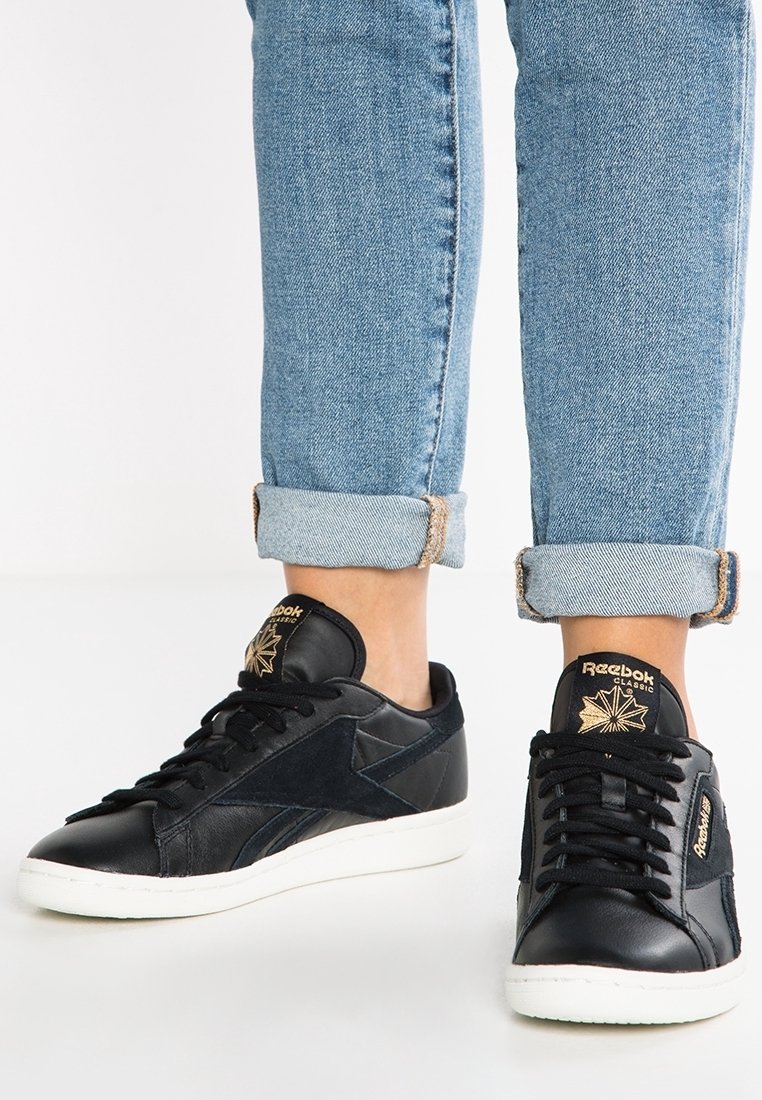 Baskets Reebok noires avec des accents en cuir texturé et en suède. Semelles en caoutchouc blanches et logo doré sur la languette. Portées avec un jean bleu retroussé.