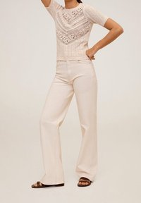 Pletený svetřík s krátkým rukávem v světlé béžové barvě s krajkovými vzory a žebrovaným lemem, spárovaný s krémovými džínami s vysokým pasem a širokými nohavicemi a hnědými sandály.