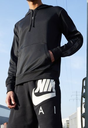 Zwarte hoodie met een gladde textuur en een kangoeroezak. Heeft een subtiele Nike-logo op de borst en patroonaccenten op de mouwen. Zwarte shorts met een groot wit Nike-logo.