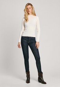 Bílý žebrovaný top s dlouhým rukávem a vysokým límcem, spojený s tmavomodrými skinny džíny a hnědými kotníkovými botami. Jednoduchý, přiléhavý design.