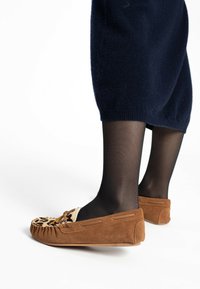 Braune Wildleder-Mokassins mit einem hellen, gemusterten Obermaterial, einer abgerundeten Zehe und dekorativen Schnürsenkeln, vor einem schlichten Hintergrund.