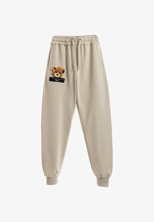 Pantalones de chándal beige con cintura elástica, que cuentan con un parche de oso texturizado en el bolsillo izquierdo y un acento de etiqueta negra encima de este.