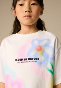 T-shirt bianco in cotone con grafica floreale colorata e testo nero che recita "FLORESCI NELLA NATURA TROVAMI NEI FIORI." Maniche corte.