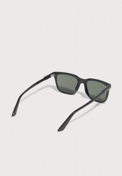 Lunettes de soleil rectangulaires noires avec verres teintés foncés, posées ouvertes sur une surface claire, montrant la charnière intérieure et un détail de marque subtil sur la branche.