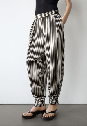 Persona che indossa pantaloni larghi a righe verticali bianche e nere con risvolti alle caviglie e sandali neri con tacco e punta aperta, in piedi contro un muro bianco.
