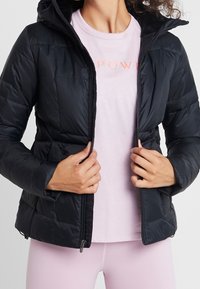 Czarna, puchowa kurtka z kapturem, zapinana na suwak z elastycznymi mankietami, noszona na jasnoróżowej koszuli z napisem "POWER" w pomarańczowym kolorze.