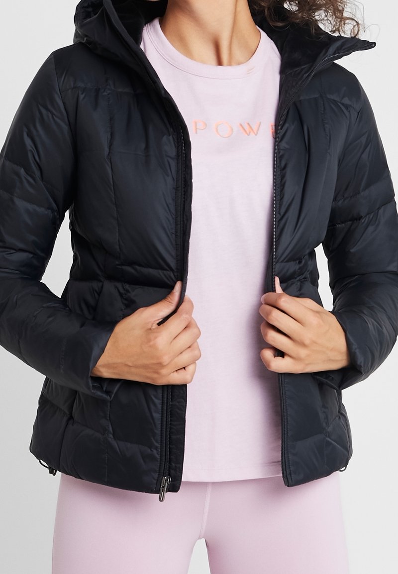 Czarna, puchowa kurtka z kapturem, zapinana na suwak z elastycznymi mankietami, noszona na jasnoróżowej koszuli z napisem "POWER" w pomarańczowym kolorze.