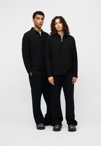 Μαύρα πουλόβερ με φερμουάρ στο λαιμό και μακριά μανίκια, συνδυασμένα με φαρδιά μαύρα παντελόνια. Το ελαφρύ ύφασμα έχει ομαλή υφή.