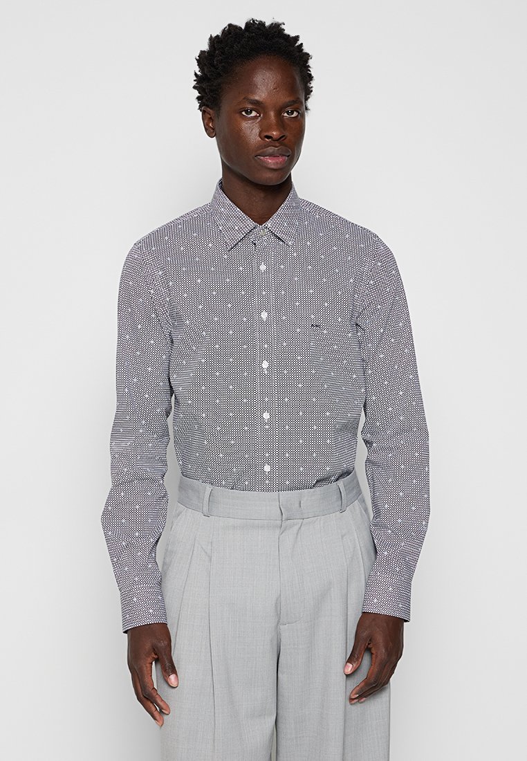 Camisa de hombre abotonada en blanco y azul marino con lunares, de mangas largas y cuello clásico. Se lleva con pantalones de pinzas en gris claro.