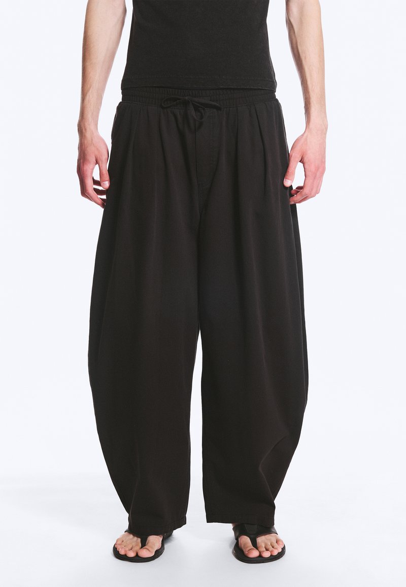 Personne portant un pantalon large noir ample avec taille à cordon et des sandales noires, debout sur fond blanc.