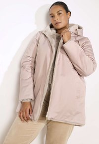 Cappotto imbottito rosa chiaro con interno in crema morbido, cappuccio con cordino e zip dorata. Presenta tasche laterali e polsini a costine.
