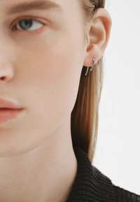 Pendientes de aro plateados con un diseño curvado, que presentan pequeñas piedras preciosas azules. El modelo tiene el cabello claro y lleva una blusa negra texturizada.