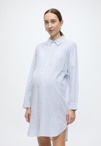 Lichtblauwe gestreepte jurk met knopen, lange mouwen, een kraag en zijstrikjes. De stof lijkt lichtgewicht en licht doorschijnend.