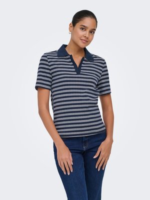 Vrouw die een marineblauw-wit gestreept polohemd met korte mouwen en een blauwe spijkerbroek draagt, staand met de handen voor zich, tegen een effen achtergrond.