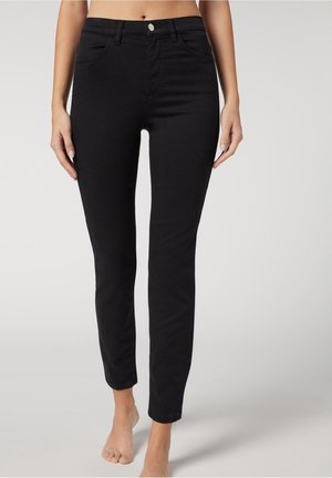 Woman wearing fitted black ankle-length jeans, standing barefoot against a plain light background.