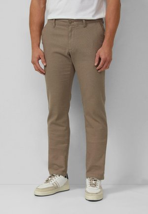 Mann trägt beiger strukturierter Hose, weißes T-Shirt und weiße Turnschuhe und steht vor einem schlichten grauen Hintergrund.