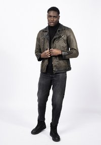 Mężczyzna ubrany w brązową skórzaną kurtkę, czarny golf, ciemnoszare jeansy i czarne buty, stojący na tle jednolitego białego tła.