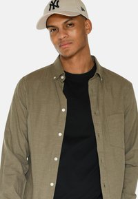 Camicia a bottone verde oliva con tasca, indossata sopra una maglietta nera. La persona indossa un cappellino beige con un logo nero.