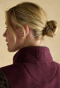 Burgunderfarbene Fleecejacke mit hohem Kragen, versehen mit dem geprägten "JOULES"-Logo, kontrastierenden braunen Akzenten und samtiger Textur.